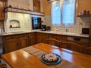 Keittiössä on puinen pöytä, sinisellä lautasella kakku, jääkaappi, kahvinkeitin ja ikkuna, jossa on pitsiverhot.