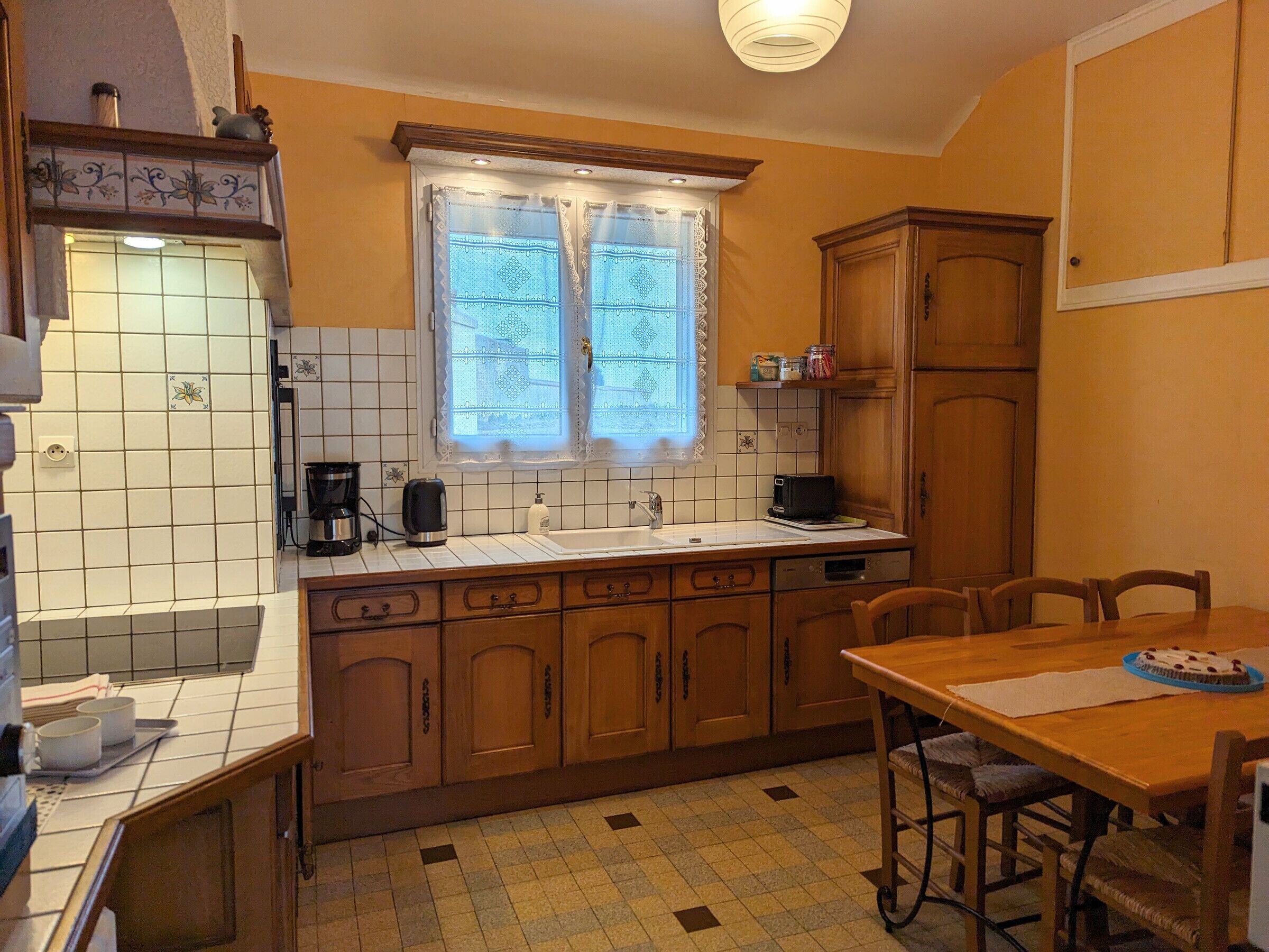 A compact kitchen with a tiled backsplash, a window with lace curtains, wooden cabinets, and a dining area with a table and chairs.