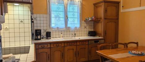 A compact kitchen with a tiled backsplash, a window with lace curtains, wooden cabinets, and a dining area with a table and chairs.