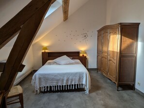A bedroom with a sloped ceiling, a wooden headboard, a bed with a white blanket, two bedside lamps, a wooden wardrobe, and a chair. - Farmhouse Retreat in Scenic Gâtinais - Spacious, Accessible & Eco-Friendly (Chailly-en-Gâtinais)