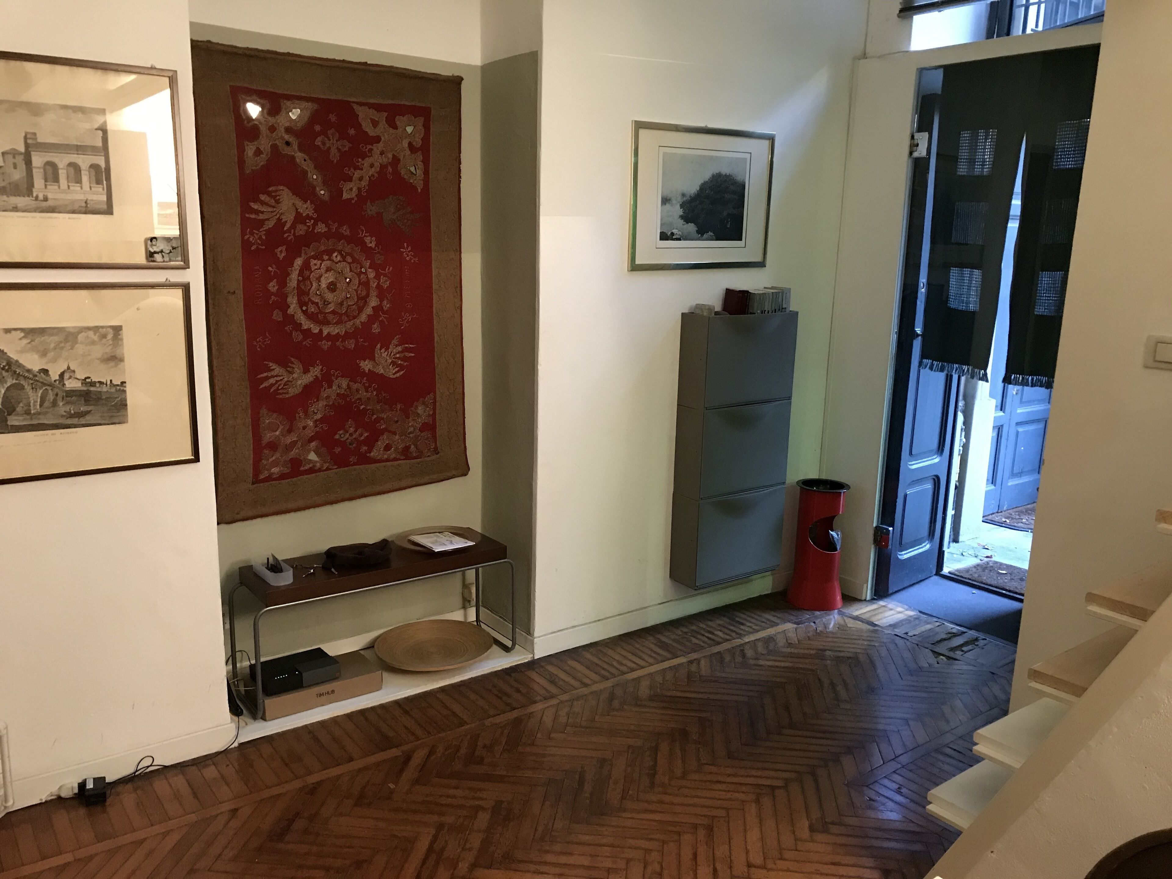 A hallway with a wooden herringbone floor, a red patterned rug, a metal shelf with a bowl, and framed pictures on the walls.