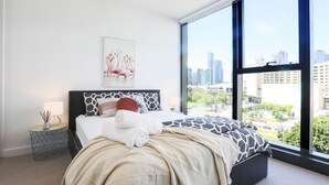 A modern bedroom with a large window, a city view, a neatly made bed, and a small bedside table with a lamp.