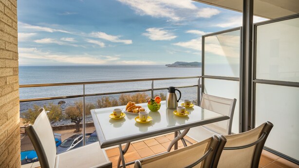 A balcony with a table set for breakfast, overlooking a body of water and a distant landmass. - Alba: Apartment on the seafront with views (L'Escala)