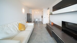 A modern living room with a white sofa, a flat-screen TV, and a dining area in the background. - Alba: Apartment on the seafront with views (L'Escala)