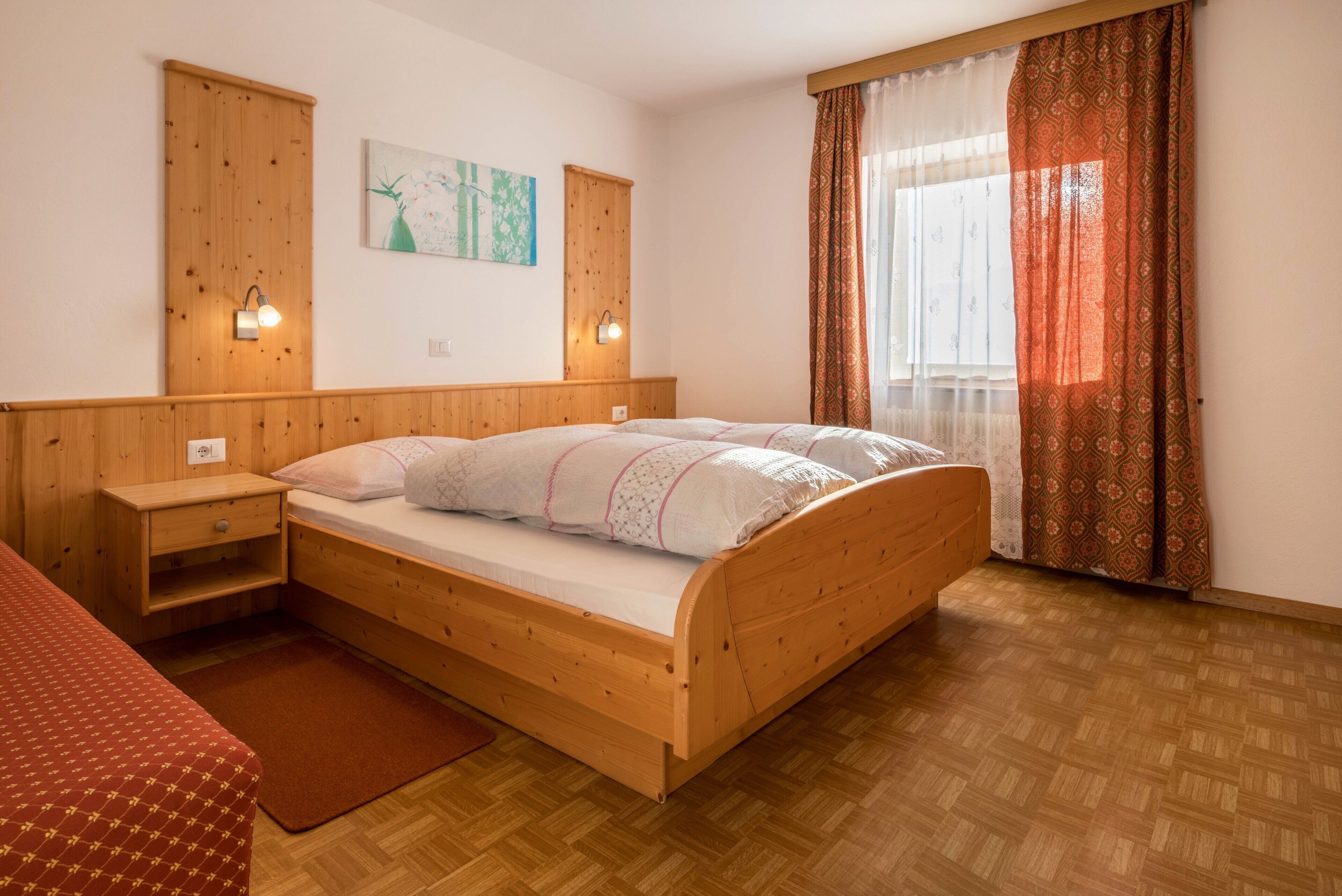 A wooden bed with a headboard, a nightstand, a red bedspread, a wooden floor, a window with patterned curtains, and a wall-mounted light fixture.