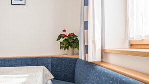 Une pièce avec un canapé bleu à motifs, une petite table recouverte d’une nappe blanche, une plante en pot avec des fleurs rouges et une fenêtre ornée de rideaux blancs.