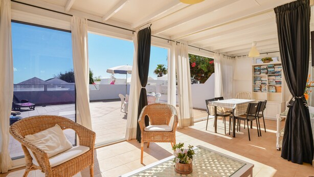 A spacious living area with a glass table, wicker chairs, a dining table, and a view through large windows.