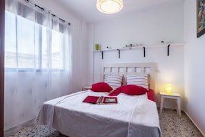 A bedroom with a bed, a bedside table with a lamp, a window with sheer curtains, and a shelf above the bed.