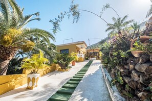 A pathway leading to a yellow building with a staircase, surrounded by palm trees and potted plants. - Holiday Home ‘Finca Alejandrina’ with Sea View, Pool, Garden & Wi-Fi (La Guancha)