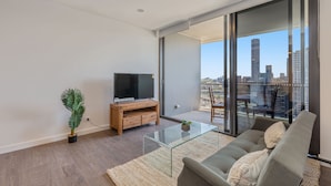 A modern living room with a glass coffee table, a sofa, a TV on a wooden stand, and a large window offering a city view. - KOZYGURU BRISBANE ONE 1Bed APT Parking (South Brisbane)