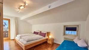 A bedroom with a wooden bed, a nightstand, a window with blinds, and a view of the outside. - Comfortable three-room vacation apartment with kitchen and south-facing balcony (Reit im Winkl)
