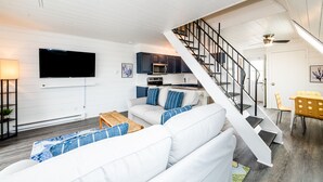 Un saló modern amb un sofà blanc, televisió de pantalla plana, zona de cuina i una escala.