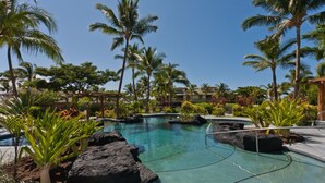 Una piscina cristalina rodeada de palmeras y rocas, con vistas a un edificio de resort al fondo.