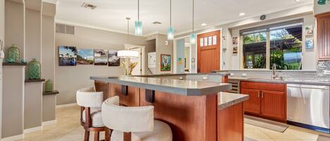 A modern kitchen with a central island, bar stools, stainless steel appliances, and a large window offering a view of greenery.