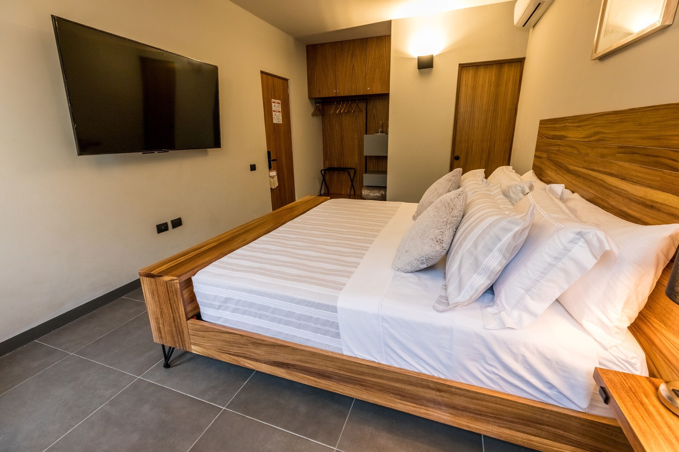 A neatly made bed with white linens and pillows, a wooden headboard, a mounted TV, and a wooden wardrobe with a mirror.
