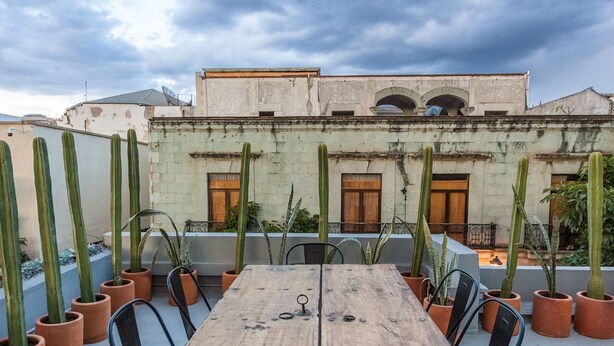 A wooden table with metal railings, surrounded by chairs, set on a rooftop terrace with potted cacti and a building in the background. - Casa Olié (Oaxaca)