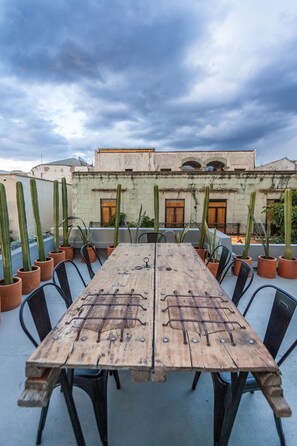 A wooden table with metal railings, surrounded by chairs, set on a rooftop terrace with potted cacti and a building in the background. - Casa Olié (Oaxaca)