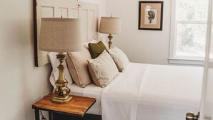 A bedroom with a bed, bedside lamps, a nightstand, a waste basket, and a window with a view of trees.