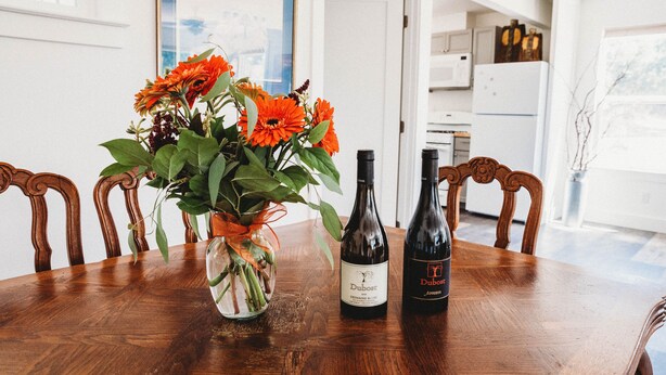 A wooden dining table with a vase of orange flowers, two wine bottles, and a glass vase with green stems.