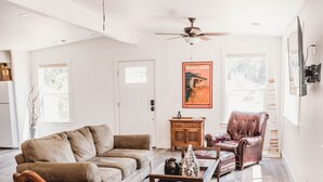 A spacious living room with a brown leather sofa, two wooden chairs with green cushions, a wooden coffee table, and a ceiling fan.