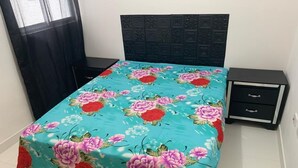 A bedroom with a bed covered in a floral bedspread, a dark headboard, a nightstand with drawers, and a window with a black curtain.