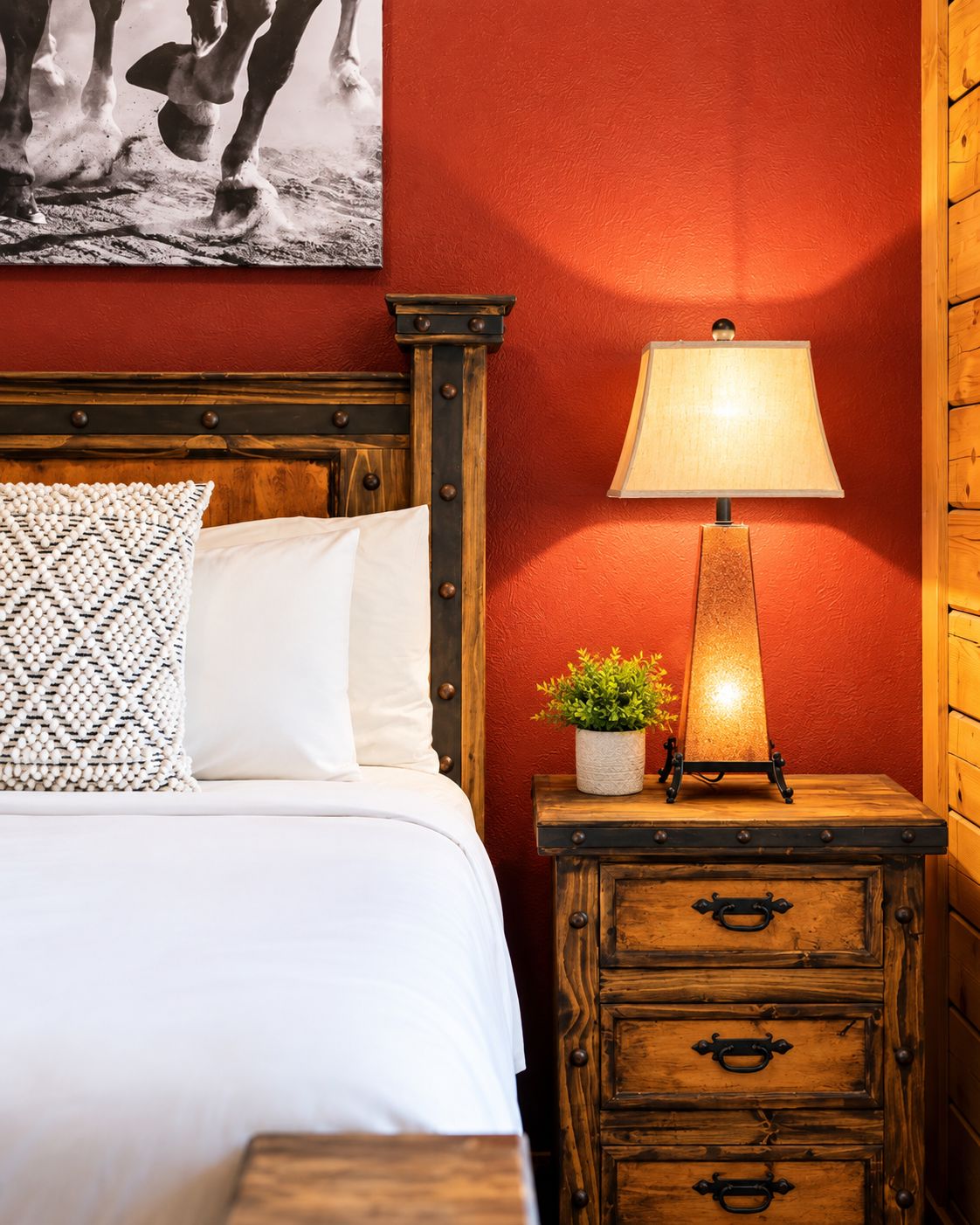 A bed with white bedding, a patterned pillow, a wooden nightstand with a lamp, and a small plant.