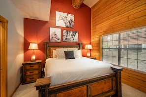 A bedroom with a wooden bed frame, two bedside tables with lamps, a red accent wall, and three framed pictures above the bed.