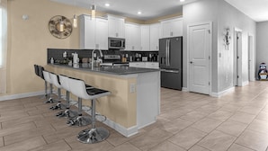 A modern kitchen with white cabinets, a central island with bar stools, stainless steel appliances, and a decorative wall piece. - Dream Vacation Getaway next to Disney with Private Pool --10BD/5BA (Kissimmee)