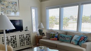 A living room with a beige sofa, a glass coffee table, a beige armchair, a TV on a cabinet, and a patterned rug. - Howards Haven (Bald Head Island)