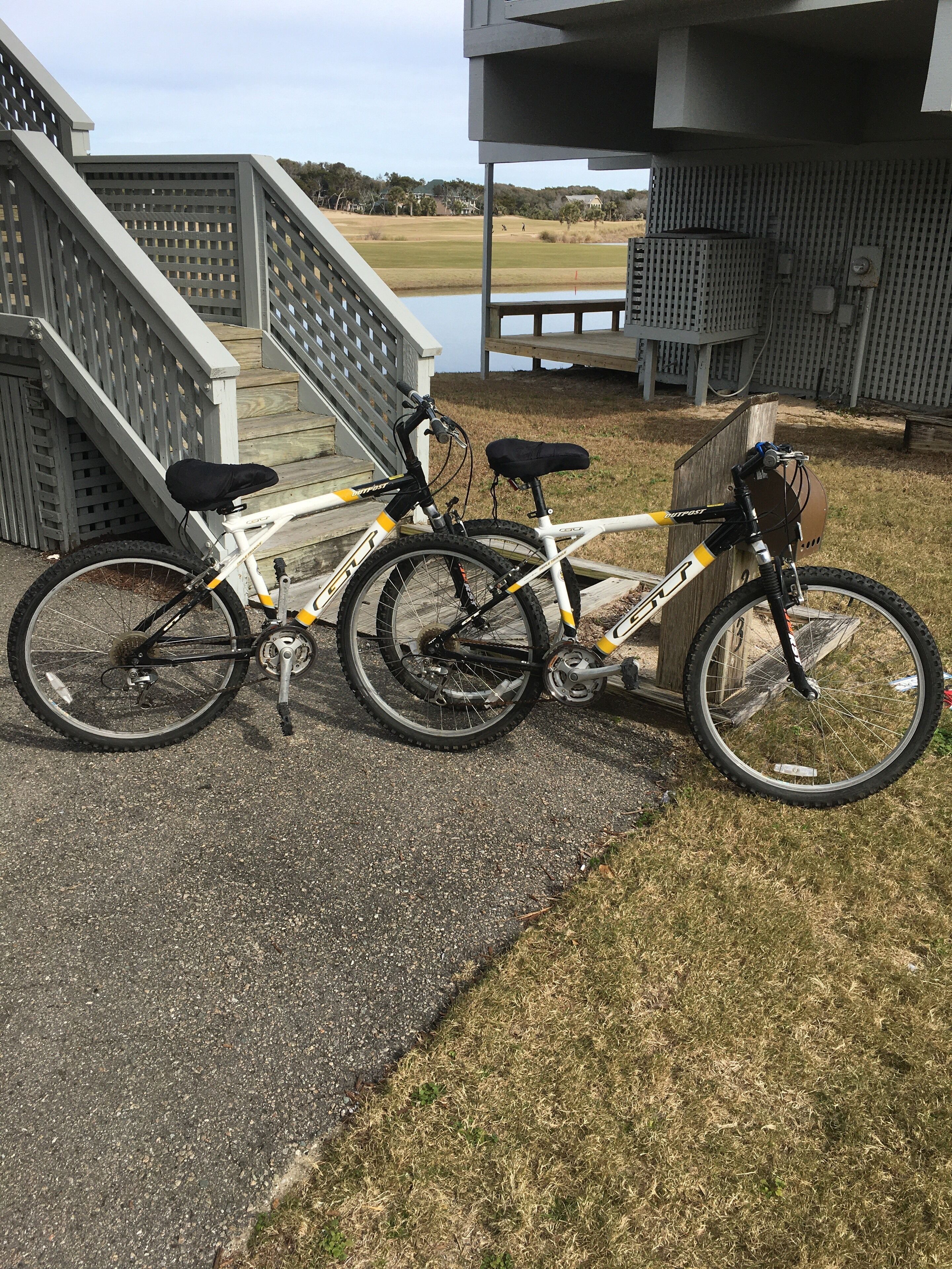 En hvid mountainbike med sorte og gule detaljer står parkeret på en belagt flade ved siden af en træterrasse og trapper, der fører op til en bygning.