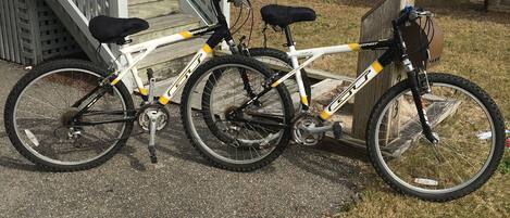 En hvid mountainbike med sorte og gule detaljer står parkeret på en belagt flade ved siden af en træterrasse og trapper, der fører op til en bygning.