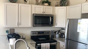 A kitchen with white cabinets, a stainless steel microwave, oven, and refrigerator. There is a sink with a faucet, and a countertop with a blender and a kettle. - Howards Haven (Bald Head Island)