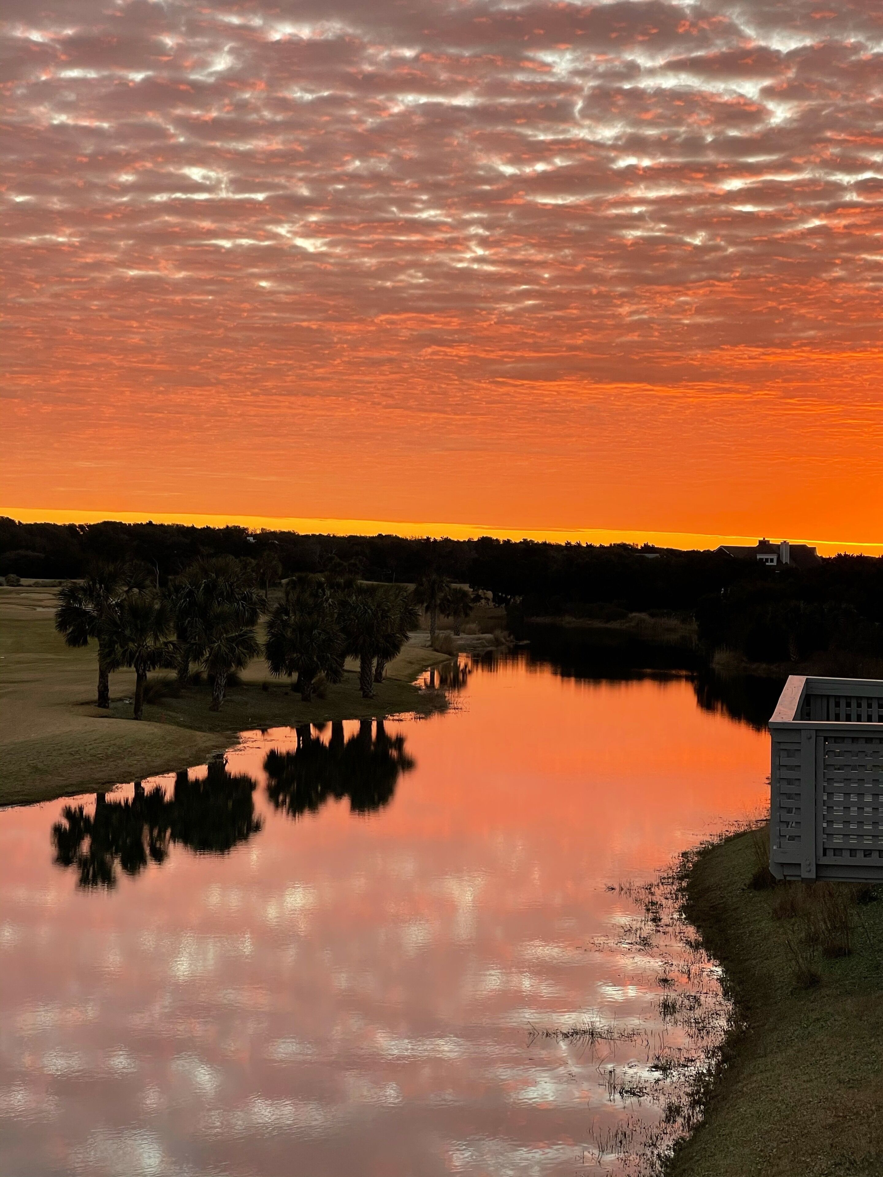 A serene sunset over a calm lake with trees and a building reflecting on the water.