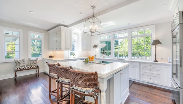 A spacious kitchen with a large island, white cabinetry, and a view of greenery through large windows.