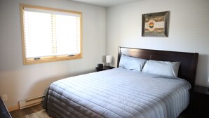 Un dormitorio con cama, mesita de noche, un reloj de pared, una ventana con persianas y un cuadro en la pared.