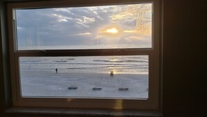 Vista desde una ventana a una playa al atardecer, con una persona caminando y tres tumbonas en la arena.