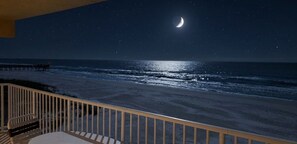 Ein Nachtblick auf einen Strand mit einem sichelförmigen Mond, einer Pier im Hintergrund und einem Balkon mit Tisch und Stuhl.
