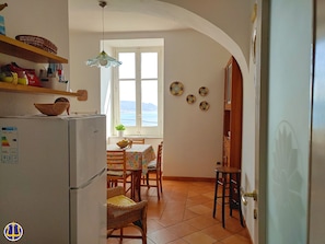 A kitchen with a refrigerator, wicker chairs, a dining table with a floral tablecloth, and a view through the window. - Villa Neptune at the beginning of the Amalfi Coast CIN No. IT065157B4VGM390GL. (Raito)