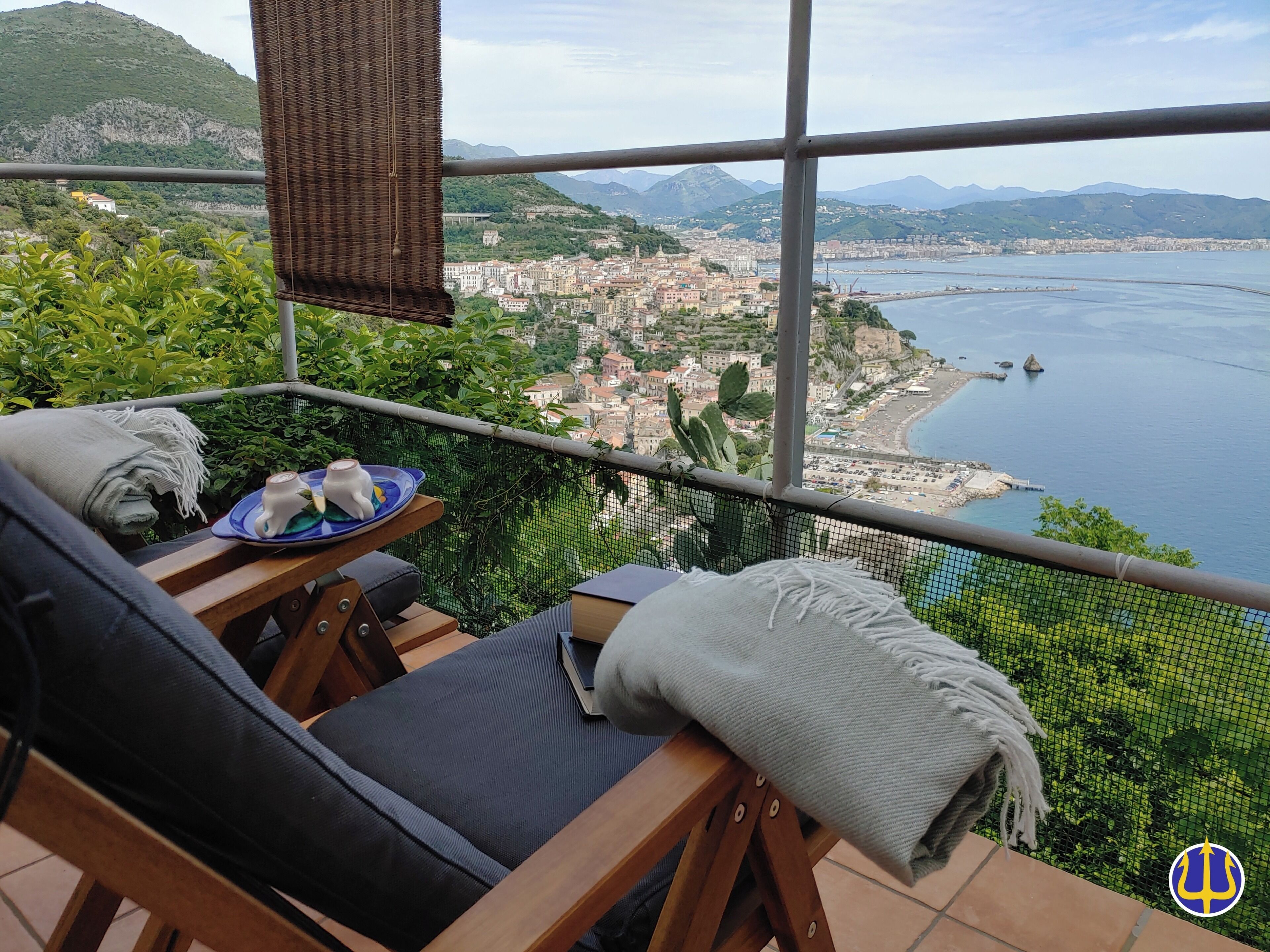 Un balcone con vista su una città costiera, un tavolo con un piatto e una tazza, e una sedia con una coperta.