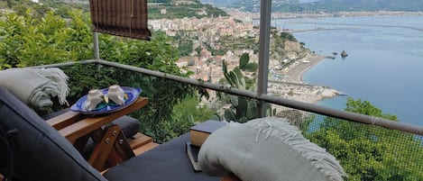 Een balkon met uitzicht op een kuststad, een tafel met een bord en een kop, en een stoel met een deken.