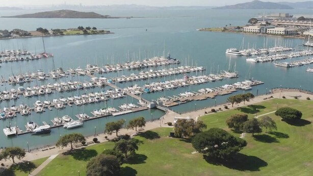 A marina with numerous boats docked in a well-organized layout, surrounded by green areas and a distant view of a cityscape. - Marina Bay Saddle (Richmond)