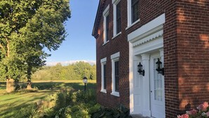 A brick house with a white door and windows, surrounded by lush greenery and a clear sky. - Majestic House on 23 acres with 5k feet of lakefront  (Fairfax)