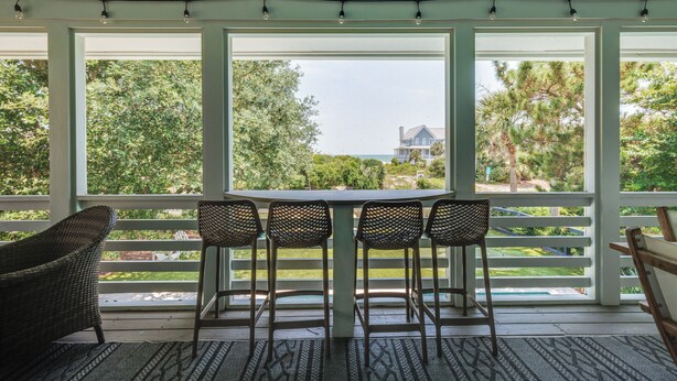 Een overdekt terras met een tafel en stoelen, grote ramen en uitzicht op groen en een huis.