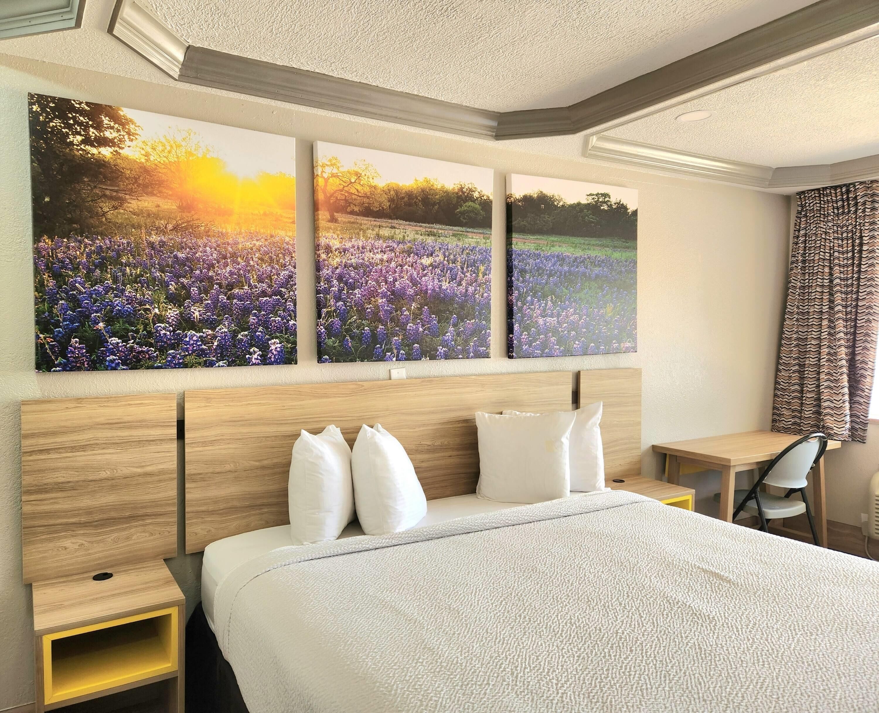 A hotel room with a large bed, a wooden headboard, a desk with a chair, and three wall-mounted paintings of a field at sunset.