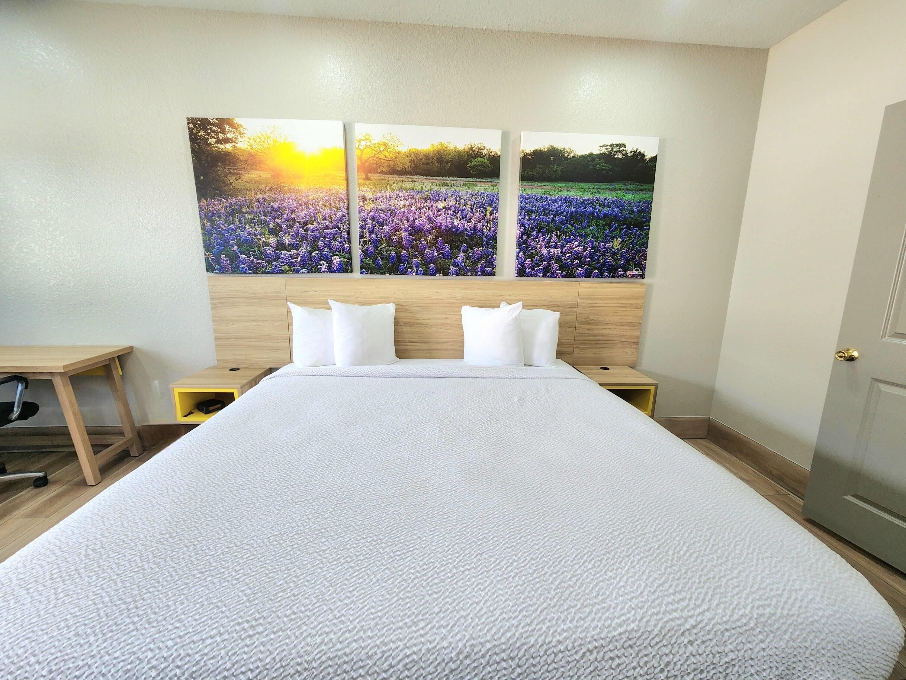 A hotel room with a large bed, two nightstands, a wooden headboard, and three wall-mounted paintings of a field with purple flowers.