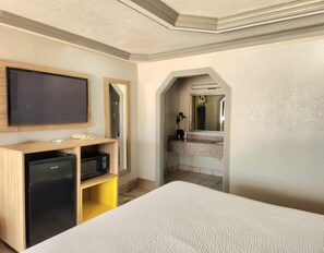 A hotel room with a bed, a TV, a desk with a computer, a mirror, and a bathroom visible through an archway.