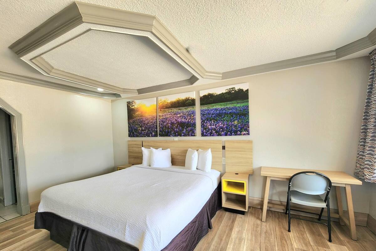A hotel room with a large bed, a desk with a chair, and a wall-mounted painting of a field.