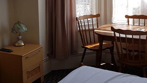 A hotel room with a bed, a wooden nightstand with a lamp, a wooden table with chairs, and a window with curtains. - Glen Villa Flat 2 - 6 Berth (Great Yarmouth)
