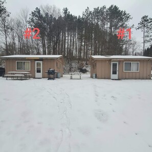 Two small cabins with a snow-covered ground, a table, and a grill outside.