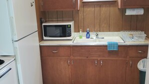 A compact kitchen with wooden cabinets, a white refrigerator, a microwave, a sink with a soap dispenser, and a window with a floral curtain. - Week/ Weekend Cabin Rental #2 (Luther)